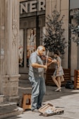 An older man is playing a violin on a street corner near a building with ornate columns. He has a violin case open on the ground in front of him, likely for collecting tips. In the background, there are posters of models on the windows of a store and a woman walking past holding a bag.