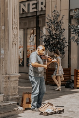 An older man is playing a violin on a street corner near a building with ornate columns. He has a violin case open on the ground in front of him, likely for collecting tips. In the background, there are posters of models on the windows of a store and a woman walking past holding a bag.