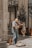 An older man is playing a violin on a street corner near a building with ornate columns. He has a violin case open on the ground in front of him, likely for collecting tips. In the background, there are posters of models on the windows of a store and a woman walking past holding a bag.