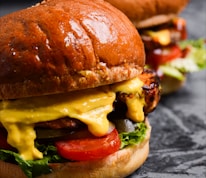 A close-up view of two juicy burgers with glossy, rich brown buns. The burgers are filled with vibrant ingredients including melted cheese, fresh lettuce, sliced tomatoes, and grilled patties.