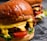 A close-up view of two juicy burgers with glossy, rich brown buns. The burgers are filled with vibrant ingredients including melted cheese, fresh lettuce, sliced tomatoes, and grilled patties.