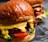 A close-up view of two juicy burgers with glossy, rich brown buns. The burgers are filled with vibrant ingredients including melted cheese, fresh lettuce, sliced tomatoes, and grilled patties.