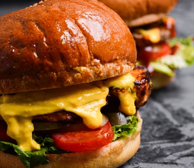 A close-up view of two juicy burgers with glossy, rich brown buns. The burgers are filled with vibrant ingredients including melted cheese, fresh lettuce, sliced tomatoes, and grilled patties.