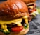 A close-up view of two juicy burgers with glossy, rich brown buns. The burgers are filled with vibrant ingredients including melted cheese, fresh lettuce, sliced tomatoes, and grilled patties.