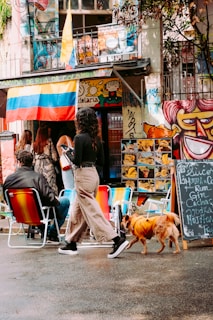 An outdoor street scene featuring several people gathered outside a vibrant, graffiti-covered building. A woman walks a small dog dressed in an orange outfit while another person sits on brightly colored outdoor chairs. A large Colombian flag hangs from the building, surrounded by colorful murals and signage with handwritten chalkboard menus displaying various drinks.