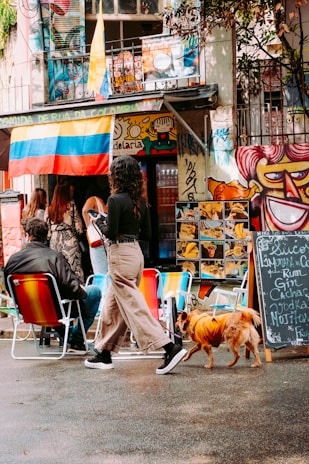 An outdoor street scene featuring several people gathered outside a vibrant, graffiti-covered building. A woman walks a small dog dressed in an orange outfit while another person sits on brightly colored outdoor chairs. A large Colombian flag hangs from the building, surrounded by colorful murals and signage with handwritten chalkboard menus displaying various drinks.