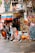An outdoor street scene featuring several people gathered outside a vibrant, graffiti-covered building. A woman walks a small dog dressed in an orange outfit while another person sits on brightly colored outdoor chairs. A large Colombian flag hangs from the building, surrounded by colorful murals and signage with handwritten chalkboard menus displaying various drinks.