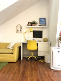 A cozy home office with a yellow theme, including a yellow chair, desk lamp, and couch. The workspace features a white desk with a shelf above it, decorated with a plant, framed quotes, and small photo frames. The floor is wooden, adding warmth to the space.