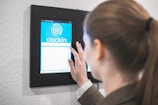 A person using a digital touch screen device mounted on a wall to clock in. The screen displays a blue interface with a circular graphic and the word 'clockin'.
