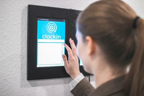 A person using a digital touch screen device mounted on a wall to clock in. The screen displays a blue interface with a circular graphic and the word 'clockin'.