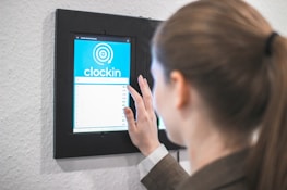 A person using a digital touch screen device mounted on a wall to clock in. The screen displays a blue interface with a circular graphic and the word 'clockin'.
