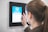 A person using a digital touch screen device mounted on a wall to clock in. The screen displays a blue interface with a circular graphic and the word 'clockin'.