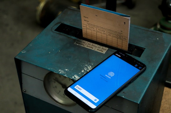 A vintage time clock machine with a paper punch card partially inserted. Next to the machine, there is a smartphone displaying a clock-in app on its screen.