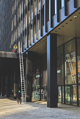 A worker in a high-visibility vest stands on a ledge of a modern building. The facade of the building has dark vertical lines which create a pattern in the architecture. There are two ladders set up against the building, one reaching up to where the worker is standing, and another person at the bottom wearing a green helmet and dark clothing.
