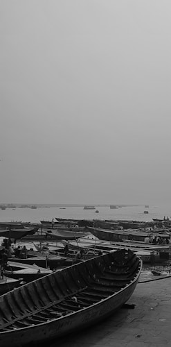 A large number of wooden boats are docked near a calm waterfront. The sky appears overcast, and the overall scene has a grayscale tone. Some people are seen around the boats, likely preparing them or engaging in other activities related to the boats.