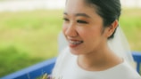 A smiling woman with braces, wearing a white outfit and holding a bouquet. She has makeup on, including eyeliner and brightly colored eyeshadow. The background is blurred, featuring greenery.