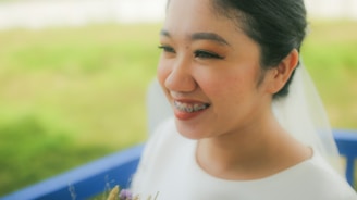 A smiling woman with braces, wearing a white outfit and holding a bouquet. She has makeup on, including eyeliner and brightly colored eyeshadow. The background is blurred, featuring greenery.