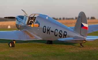 A vintage light aircraft with a sleek design is parked on a grassy field. The aircraft has a silver body with a cockpit open and a visible propeller at the front. It bears the registration number 'OK-CSR' on the side and rear, along with a small Czech flag symbol on the tail fin. The sunlight gives the body a reflective sheen, and the surrounding environment suggests an open, rural landscape.