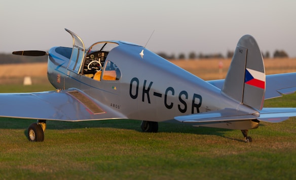 A vintage light aircraft with a sleek design is parked on a grassy field. The aircraft has a silver body with a cockpit open and a visible propeller at the front. It bears the registration number 'OK-CSR' on the side and rear, along with a small Czech flag symbol on the tail fin. The sunlight gives the body a reflective sheen, and the surrounding environment suggests an open, rural landscape.