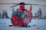 A bright red helicopter is stationary on a snowy ground surrounded by snow-covered trees in a cold, wintery landscape.