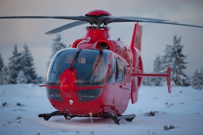 A bright red helicopter is stationary on a snowy ground surrounded by snow-covered trees in a cold, wintery landscape.