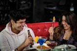 Two people are sitting on a red cushioned bench, laughing and enjoying their time together. One is holding a beer bottle, and the other is holding a fork with food. Various colorful cocktails and dishes with food are placed on the table in front of them.