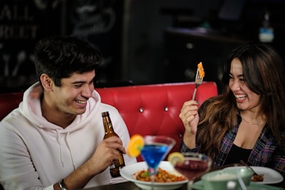 Two people are sitting on a red cushioned bench, laughing and enjoying their time together. One is holding a beer bottle, and the other is holding a fork with food. Various colorful cocktails and dishes with food are placed on the table in front of them.