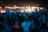 A large crowd of people is gathered at an outdoor event during the night. Many individuals are raising their hands and holding up smartphones, capturing the moment. The scene is illuminated by bright spotlights in the background and various colored lights creating a vibrant atmosphere.