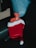 A red hot water bottle is partially tucked inside a red Santa hat with a white brim and pom-pom. The scene is set against a black leather surface and a blue cushion can be seen in the background. A TV remote control is visible next to the bottle.
