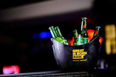 A black ice bucket containing several green beer bottles prominently displaying the brand 'Carlsberg'. The bucket has a yellow text label that reads 'GREAT IS BORN RAW.' and '#Playcraft Gi Copter7'. The background is blurred with colorful lights, suggesting it is possibly taken in a bar or club setting.