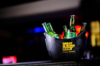 A black ice bucket containing several green beer bottles prominently displaying the brand 'Carlsberg'. The bucket has a yellow text label that reads 'GREAT IS BORN RAW.' and '#Playcraft Gi Copter7'. The background is blurred with colorful lights, suggesting it is possibly taken in a bar or club setting.