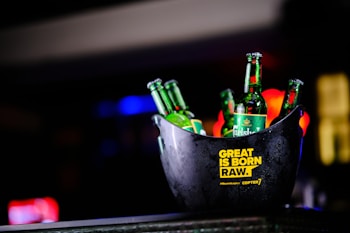 A black ice bucket containing several green beer bottles prominently displaying the brand 'Carlsberg'. The bucket has a yellow text label that reads 'GREAT IS BORN RAW.' and '#Playcraft Gi Copter7'. The background is blurred with colorful lights, suggesting it is possibly taken in a bar or club setting.