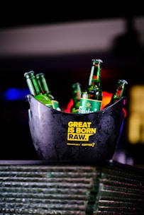 Several green Carlsberg beer bottles are placed inside a dark ice bucket. The bucket has yellow text printed on it reading 'Great is Born Raw.' The setting is dimly lit, creating a vibrant bar or party ambiance.