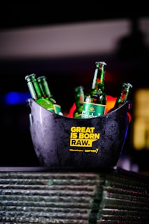 Several green Carlsberg beer bottles are placed inside a dark ice bucket. The bucket has yellow text printed on it reading 'Great is Born Raw.' The setting is dimly lit, creating a vibrant bar or party ambiance.