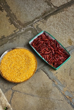 A top-down view of two containers placed on a stone floor. The container on the left holds a circular tray filled with yellow lentils, while the rectangular container on the right is filled with dried red chili peppers.