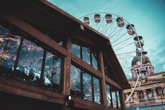 A large wooden structure with reflective glass windows displaying bright interior decorations. Adjacent to it is a Ferris wheel with several gondolas visible against a blue sky. In the background, an ornate building with a clock tower is partially visible.