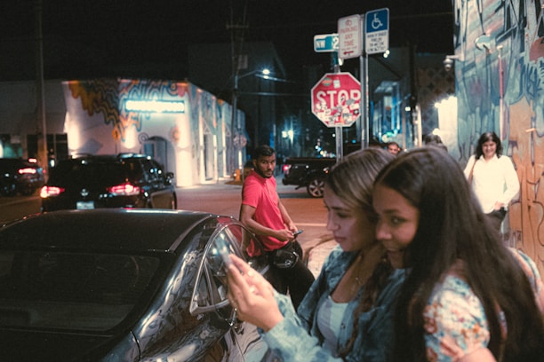 People are gathered on a dimly lit urban street at night. Two women are closely looking at a smartphone, while a man in a red shirt stands in the background looking towards them. The street is decorated with colorful murals, and several cars and street signs, including a stop sign, are visible.