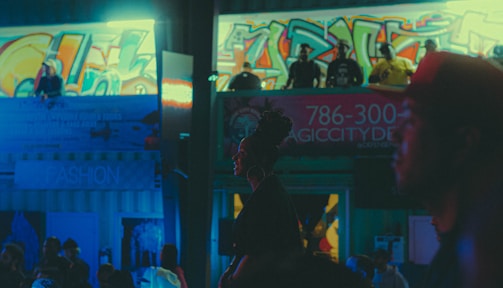 An urban night scene with vibrant graffiti covering the walls in the background. There are several people standing around and looking down from an elevated platform. The neon lights and cool blue tones dominate the atmosphere, giving it an energetic and somewhat edgy vibe. A prominent sign with a phone number and the words 'Magic City' is visible, suggesting a club or nightlife setting. The silhouettes and shadows create a moody ambiance.