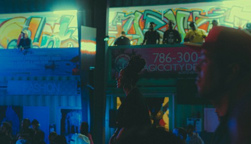 An urban night scene with vibrant graffiti covering the walls in the background. There are several people standing around and looking down from an elevated platform. The neon lights and cool blue tones dominate the atmosphere, giving it an energetic and somewhat edgy vibe. A prominent sign with a phone number and the words 'Magic City' is visible, suggesting a club or nightlife setting. The silhouettes and shadows create a moody ambiance.