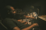 A dimly lit recording studio with two individuals seated in chairs. One person is in focus, wearing a floral shirt and a hat, while the other is blurred in the foreground. The setting includes studio equipment like audio mixers and cables.
