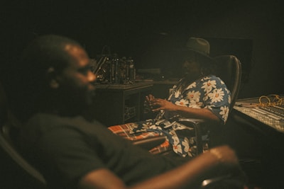 A dimly lit recording studio with two individuals seated in chairs. One person is in focus, wearing a floral shirt and a hat, while the other is blurred in the foreground. The setting includes studio equipment like audio mixers and cables.