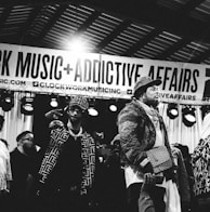 Several individuals are standing on a stage, with one person prominently in the foreground wearing a patterned jacket and a crossbody bag. There is a banner overhead with the words 'CLOCKWORK MUSIC + ADDICTIVE AFFAIRS'. Stage lights are visible in the background, and another person is holding up a mobile phone in the lower part of the image. The scene appears to be a live event or performance.