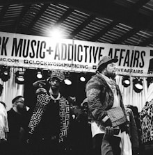 Several individuals are standing on a stage, with one person prominently in the foreground wearing a patterned jacket and a crossbody bag. There is a banner overhead with the words 'CLOCKWORK MUSIC + ADDICTIVE AFFAIRS'. Stage lights are visible in the background, and another person is holding up a mobile phone in the lower part of the image. The scene appears to be a live event or performance.