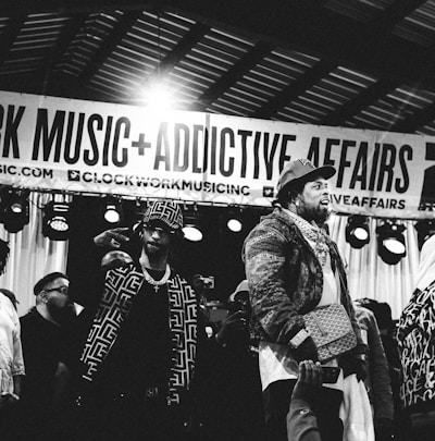 Several individuals are standing on a stage, with one person prominently in the foreground wearing a patterned jacket and a crossbody bag. There is a banner overhead with the words 'CLOCKWORK MUSIC + ADDICTIVE AFFAIRS'. Stage lights are visible in the background, and another person is holding up a mobile phone in the lower part of the image. The scene appears to be a live event or performance.