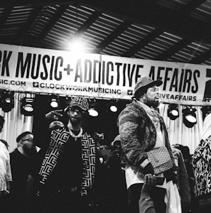 Several individuals are standing on a stage, with one person prominently in the foreground wearing a patterned jacket and a crossbody bag. There is a banner overhead with the words 'CLOCKWORK MUSIC + ADDICTIVE AFFAIRS'. Stage lights are visible in the background, and another person is holding up a mobile phone in the lower part of the image. The scene appears to be a live event or performance.