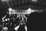 A monochrome image captures a lively musical event with a performer on stage, surrounded by a group of people. The background features a banner with the words 'CLOCKWORK MUSIC + ADDICTIVE AFFAIRS'. Several audience members hold up phones, likely recording the performance.