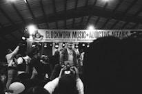 A monochrome image captures a lively musical event with a performer on stage, surrounded by a group of people. The background features a banner with the words 'CLOCKWORK MUSIC + ADDICTIVE AFFAIRS'. Several audience members hold up phones, likely recording the performance.
