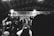 A monochrome image captures a lively musical event with a performer on stage, surrounded by a group of people. The background features a banner with the words 'CLOCKWORK MUSIC + ADDICTIVE AFFAIRS'. Several audience members hold up phones, likely recording the performance.