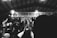 A monochrome image captures a lively musical event with a performer on stage, surrounded by a group of people. The background features a banner with the words 'CLOCKWORK MUSIC + ADDICTIVE AFFAIRS'. Several audience members hold up phones, likely recording the performance.