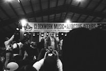 A monochrome image captures a lively musical event with a performer on stage, surrounded by a group of people. The background features a banner with the words 'CLOCKWORK MUSIC + ADDICTIVE AFFAIRS'. Several audience members hold up phones, likely recording the performance.